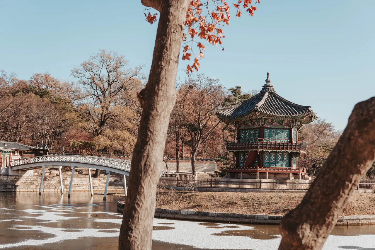 Traditional Korean pavilion by a pond, framed by trees with autumn leaves. A bridge spans the water under a clear blue sky.
