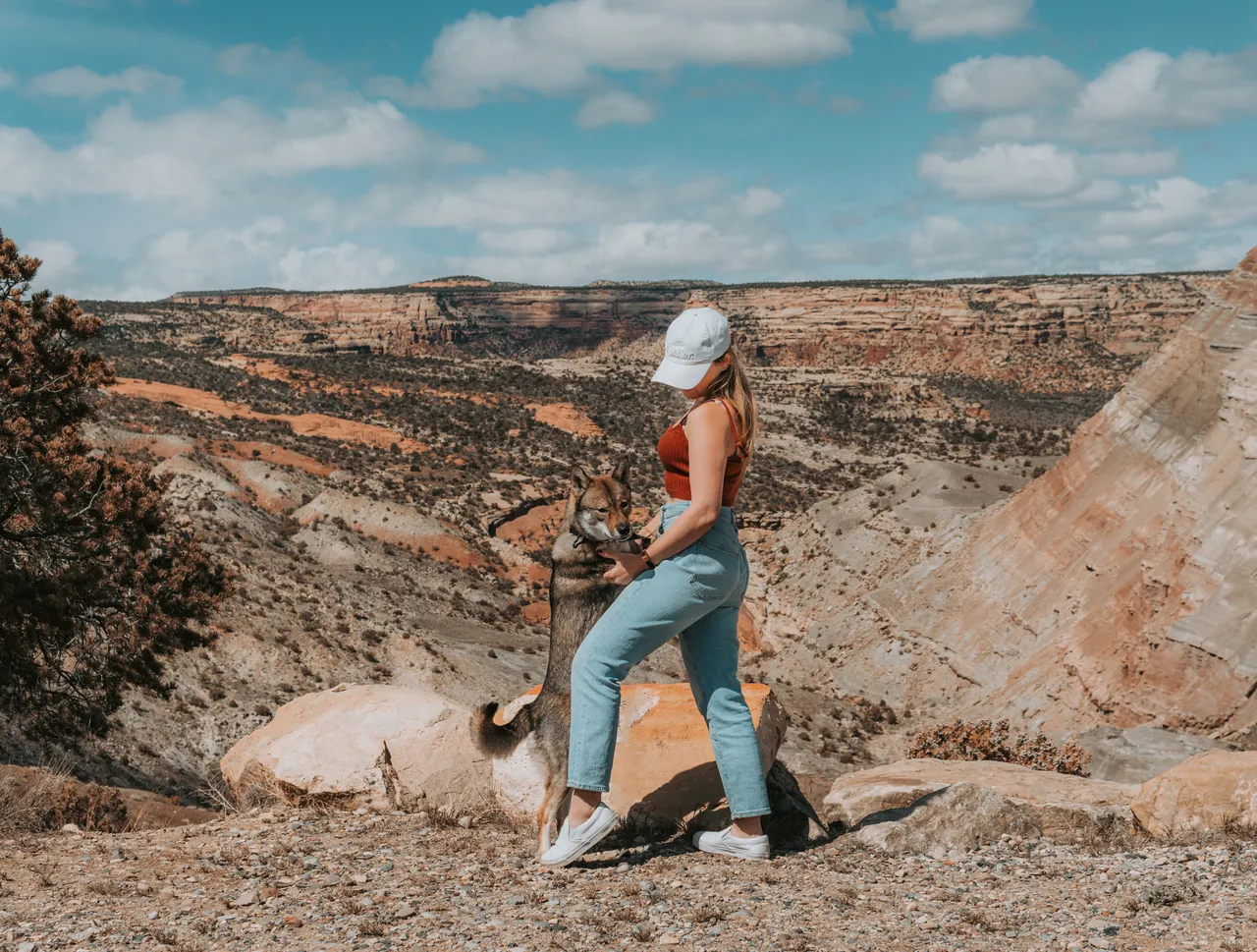 Woman in a cap hugs dog on a rocky landscape with colorful canyon in grand junction colorado. Bright day with blue sky and clouds, creating a cheerful mood.