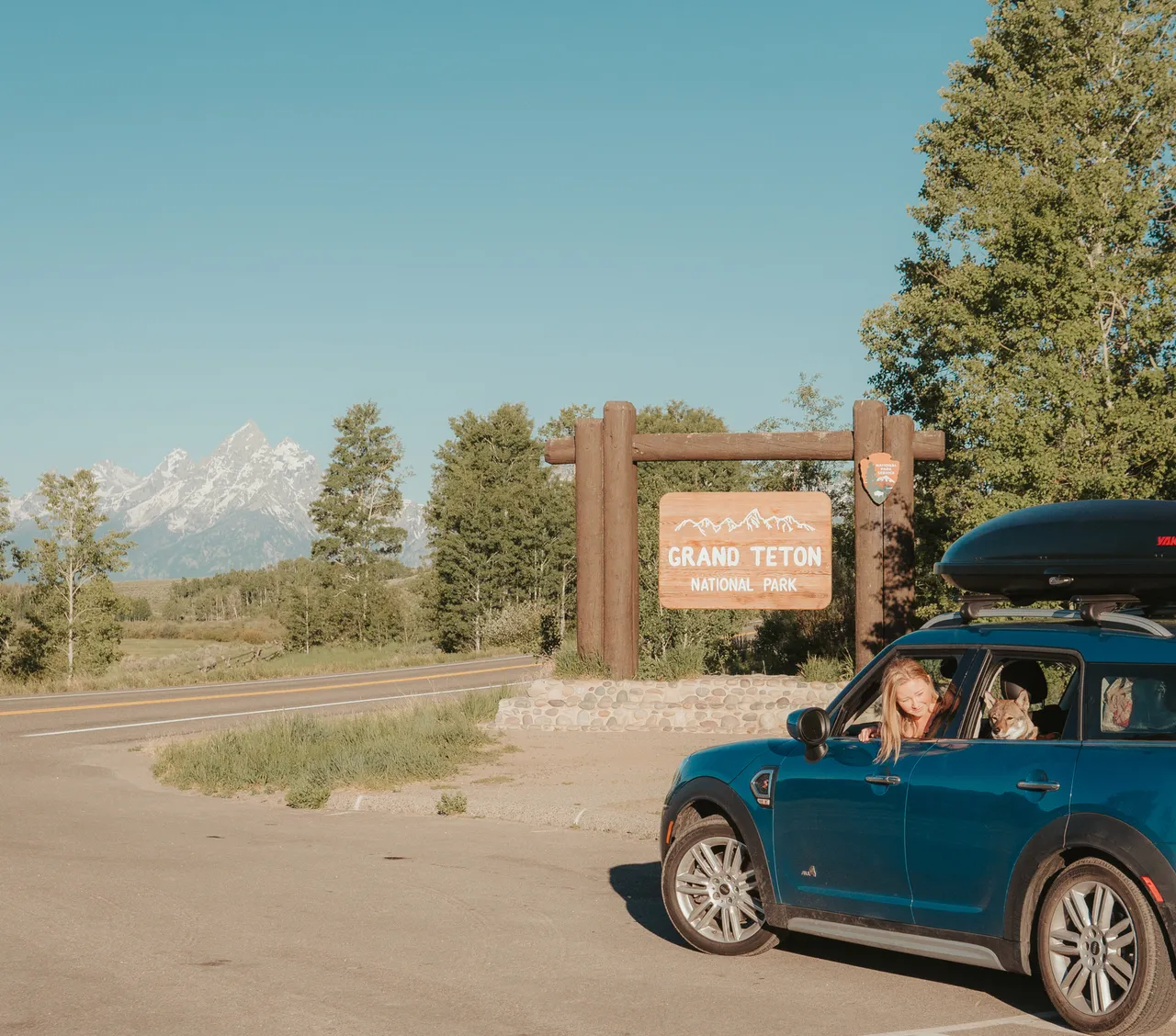 Blue MINI cooper countryman car with woman and dog at Grand Teton National Park sign. Mountain view, clear sky, and lush trees create a serene setting.