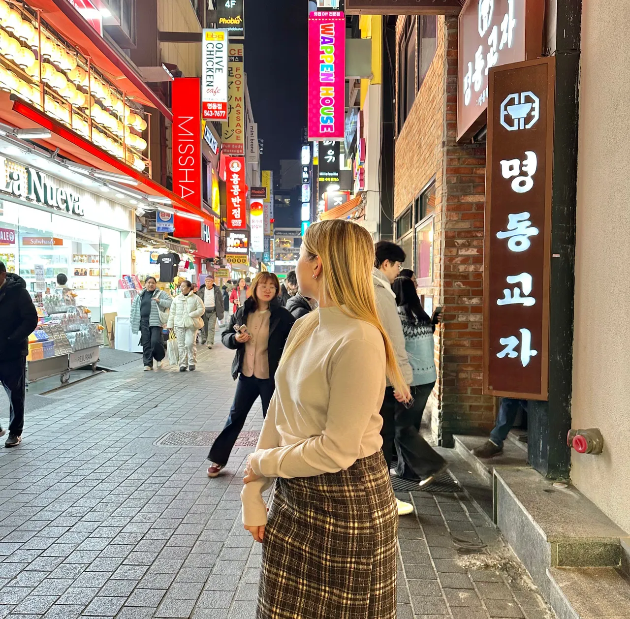 Woman in plaid skirt stands on a vibrant, busy street lined with colorful signs and bustling shoppers at night in Seoul Korea. Neon lights illuminate the scene.