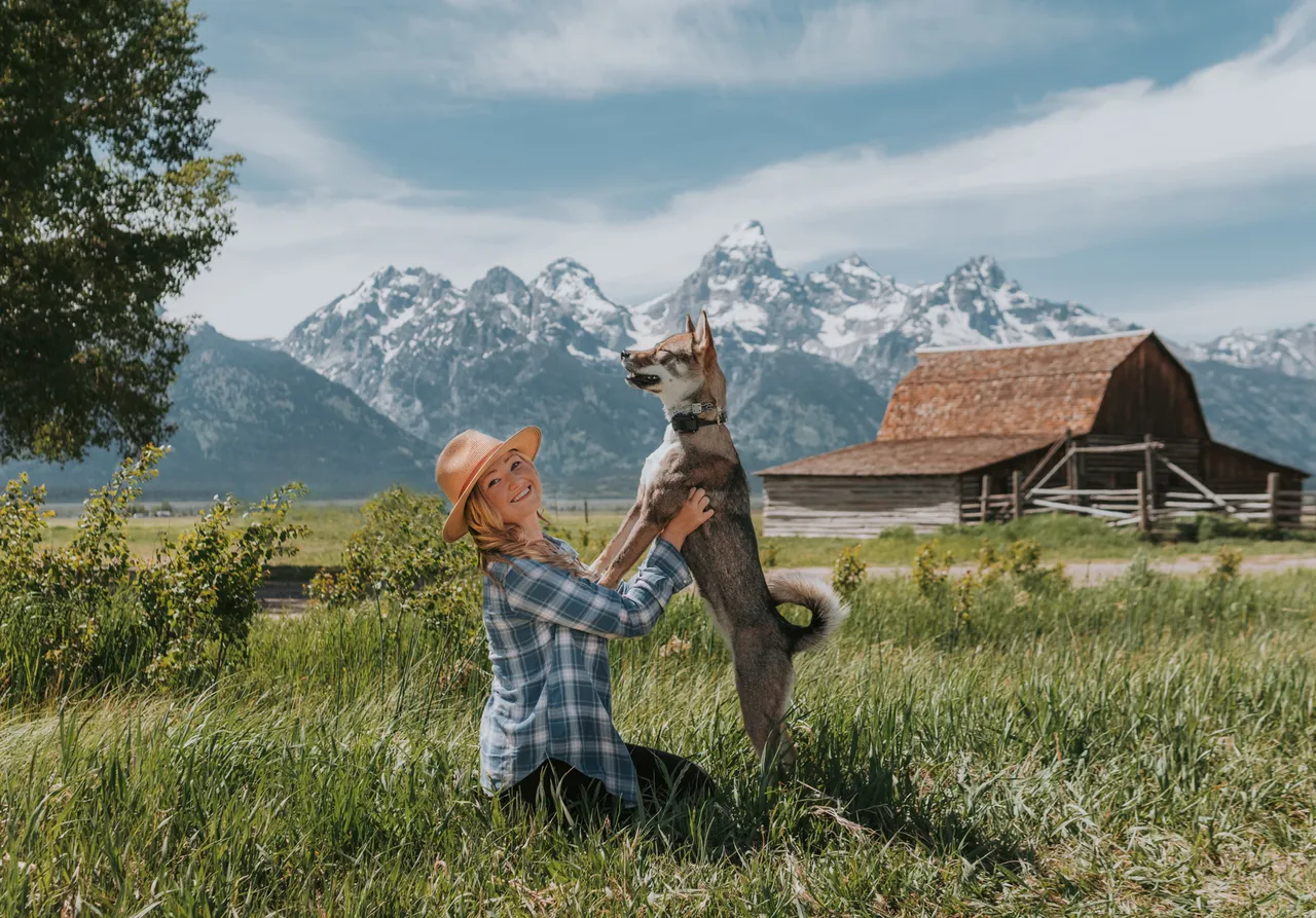 Woman in plaid shirt and hat kneels in a field, joyfully holding a standing dog. Snow-capped mountains and a barn are in the background.