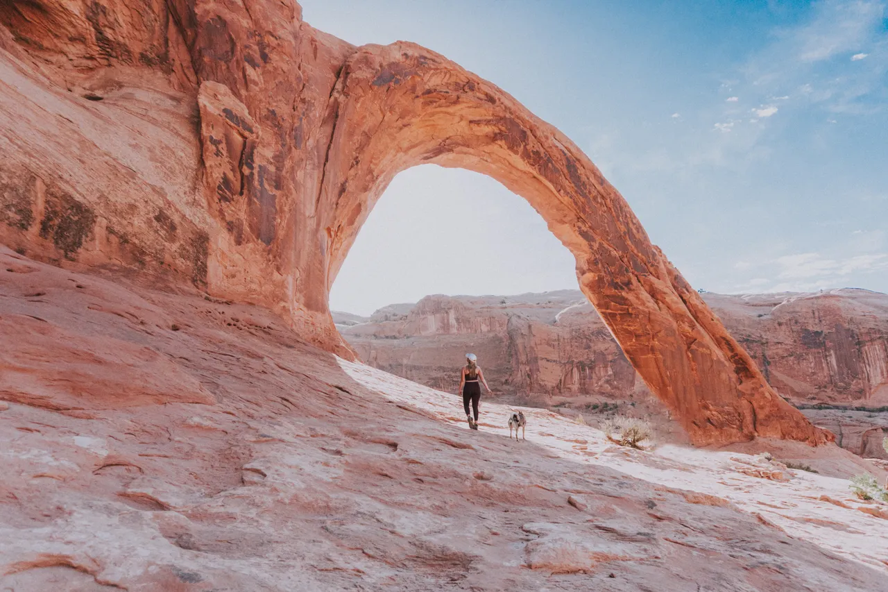 A person and dog hike under a large Corona arch in a desert landscape. Clear blue sky and rugged terrain create a serene, adventurous mood.
