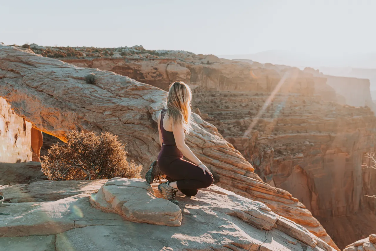 a white woman kneeling on a rock at mesa arch at sunrise with the sun shining