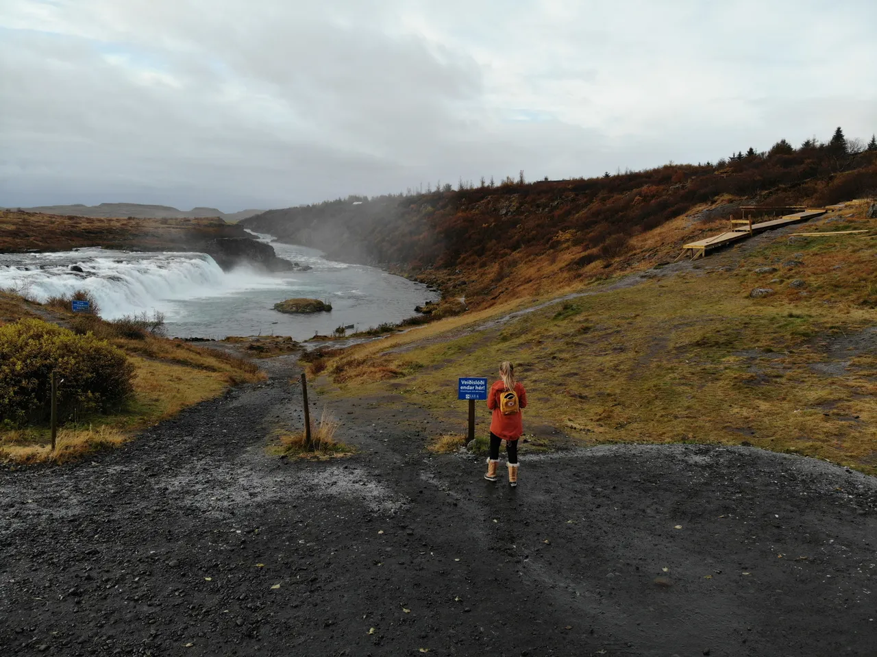Woman stands facing a waterfall with mist, wearing an orange jacket and backpack. Noticeable sign and wooden path nearby. Overcast sky.