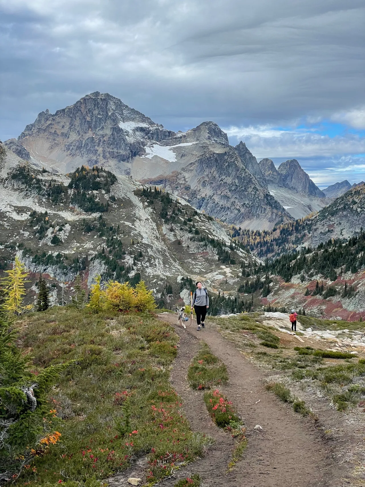 Hikers on a mountain trail below rocky peaks and a cloudy sky
