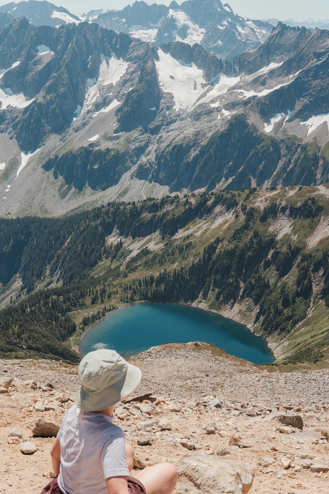 Hiker in bucket hat sitting on a ridge overlooking a turquoise alpine lake and snow-capped peaks