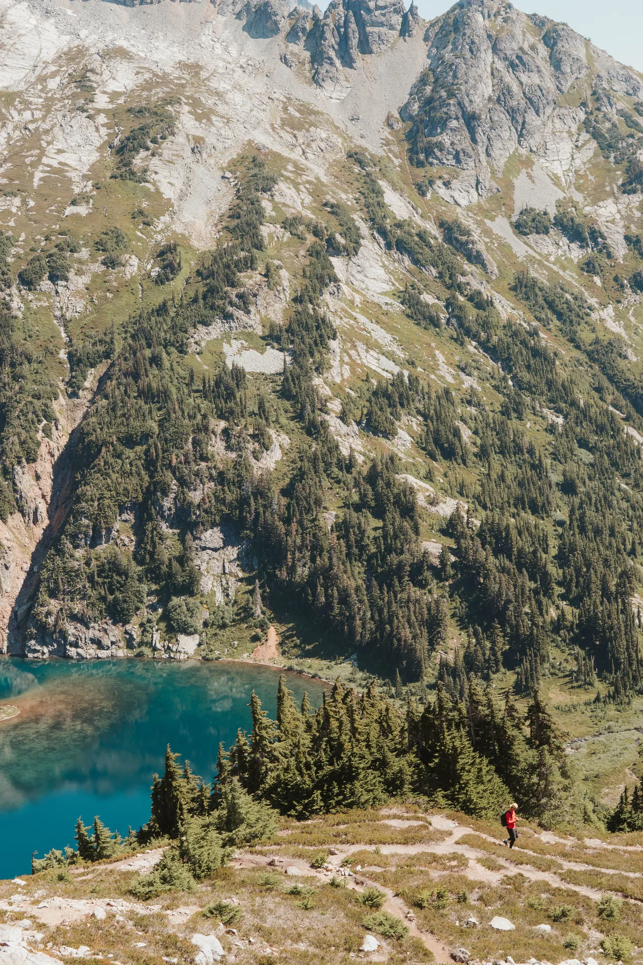Hiker on the trail above a turquoise alpine lake surrounded by steep forested mountains