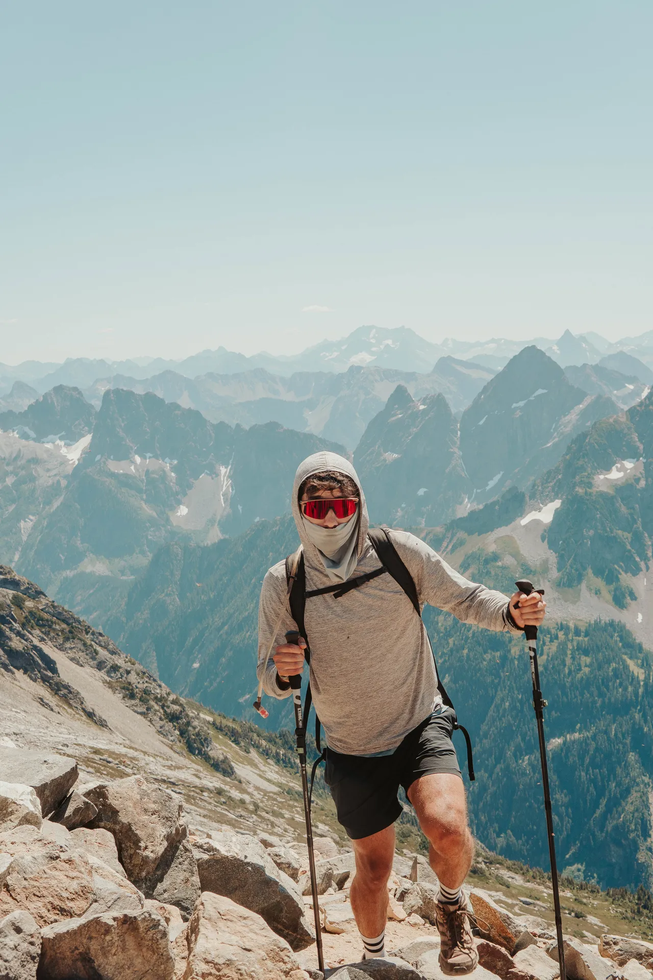 Hiker fully covered in UPF sun hoodie and sunglasses ascending Sahale Arm with North Cascades panorama behind